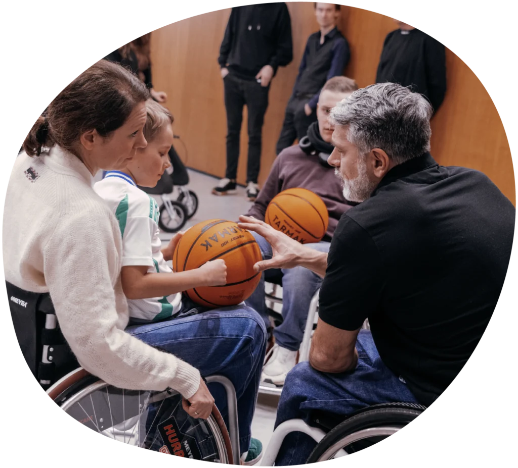 Frau mit Kind auf dem Schoß im Rollstuhl auf dem Basketballfeld bei einem Mutmacher Pioniere Workshop im Gespräch mit einem Mann im Rollstuhl